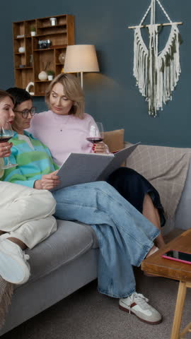 Mature Caucasian woman showing photo album to her best friends while sitting on sofa in living room
