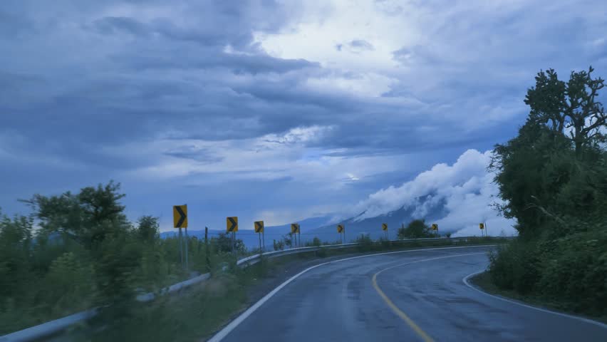 Point of View Driving Along Winding Mountain Highway Surrounded by Lush Hills, Dramatic Clouds, Scenic Blue Sky. Serene Road Trip Through Nature with Curve Warning Signs and Foggy Peaks in Distance