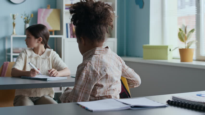 Waist up of concentrated young African American girl opening her pencil case and preparing for schoolwork, while another student writing in notebook in background of sunlit classroom