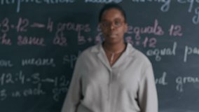 Chest up slowmo portrait of confident young African American female teacher in glasses smiling at camera while standing against classroom blackboard - Powered by Shutterstock - Get 15% off with code: PIKWIZARD15
