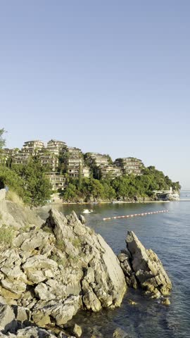 Modern Terraced Hotel Complex Built on Rocky Coastal Cliff Surrounded by Green Trees, Overlooking Calm Sea at Sunset. Distant Island, Clear Sky, and Tranquil Ocean in Scenic Mediterranean Landscape.