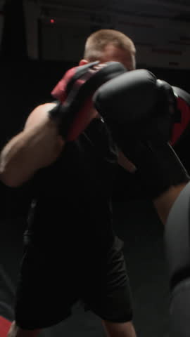 Vertical slowmo of trainer holding red mitts while boxer strikes controlled punches, refining form and timing in focused gym training