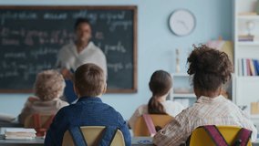 Back view shot of two engaged young students raising their hands to ask or answer during interactive Math lesson with modern Black female teacher standing near chalkboard - Powered by Shutterstock - Get 15% off with code: PIKWIZARD15