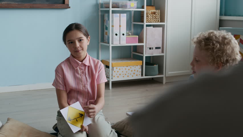 Asian schoolgirl with road sign in hands speaking about safety and awareness on road to teacher and classmates, sitting in circle on rug in bright, well-lit classroom
