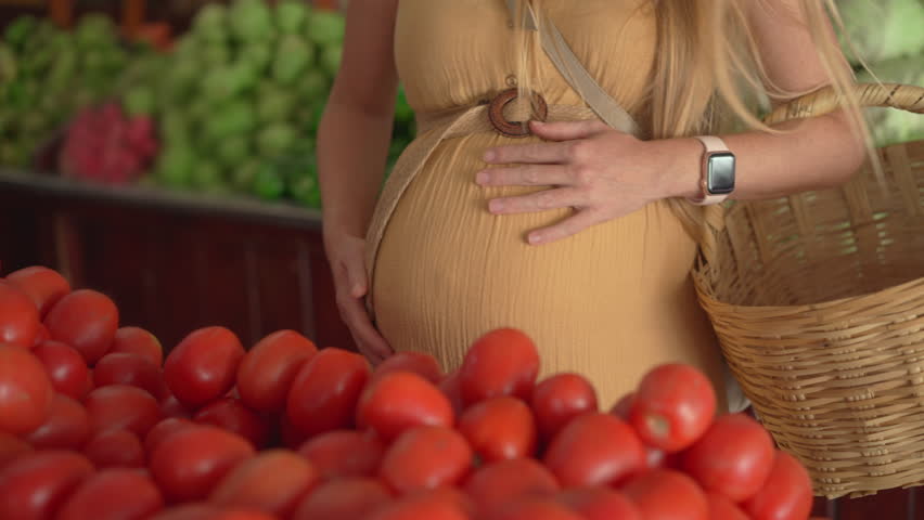 Pregnant woman in a yellow dress choosing fresh fruits, vegetables, and spices at an outdoor organic farmers market. Healthy pregnancy lifestyle, natural food, and maternal care.