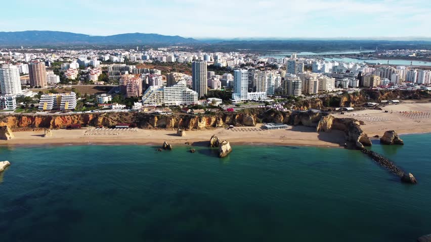 Aerial view of modern city buildings along sandy beach with rocky cliffs and calm blue sea in Faro, Portugal. Clear sky, coastal charm, and urban contrast create a vibrant scenic atmosphere.