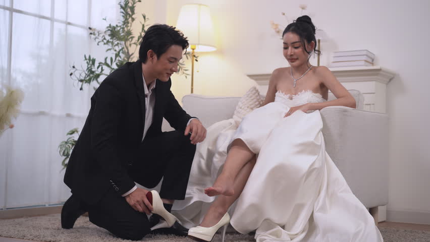 Handsome Asian man is helping a girlfriend put on her wedding shoes. The man is kneeling down and the woman is sitting on a couch. Concept of elegance and celebration.