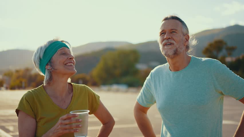 Senior couple recovering and drinking water while resting after energetic outdoor fitness session, sharing happy moment during summer workout