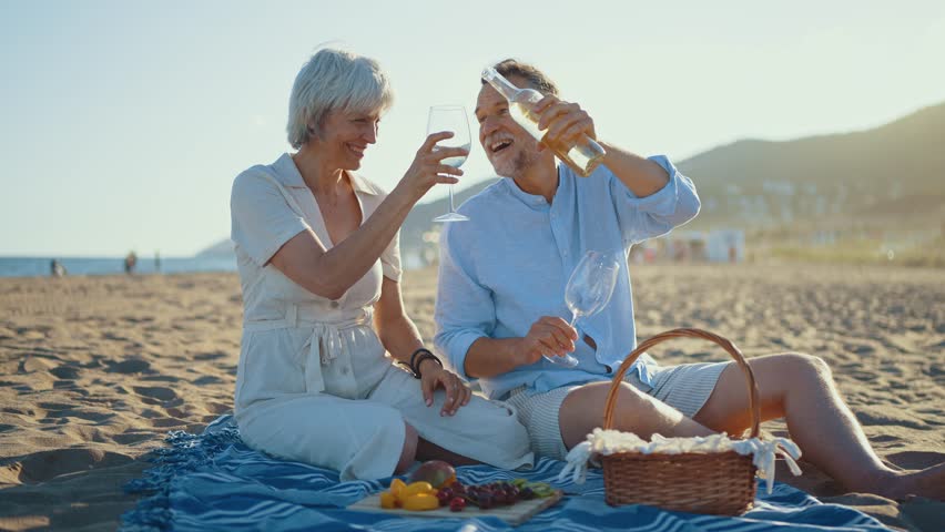 Retired couple sharing intimate moment while pouring white wine, sitting on beach blanket with picnic basket, enjoying fresh fruits during golden sunset by ocean shore