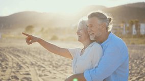 Happy retired couple enjoying a romantic moment on the beach, pointing at something in the distance while laughing together, bathed in golden sunlight. - Powered by Shutterstock - Get 15% off with code: PIKWIZARD15
