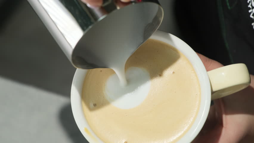 Woman slowly adds milk to coffee forming design on surface. Barista presents last phase of latte creation. Process of coffee preparation