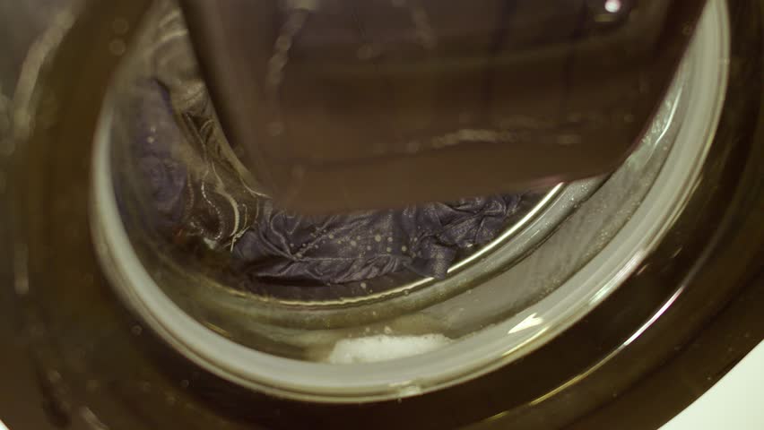 Close-up view of laundry spinning inside a front-loading washing machine. Clothes and soap suds move in slow circular motion behind the glass door.