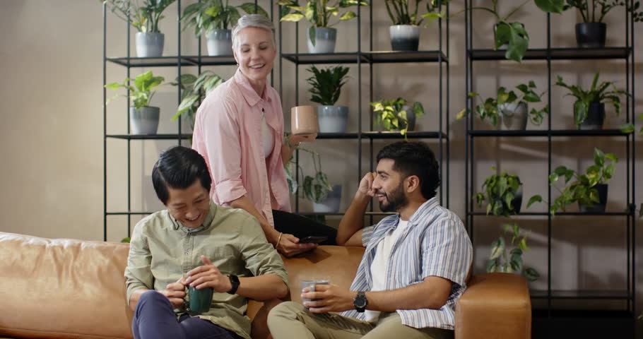 Diverse coworkers sharing drinks and laughing in office lounge after showing smartphone message. Collaboration, collegiality, camaraderie, informal, relaxation, discussion, community