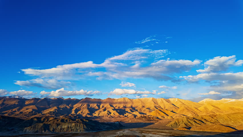 Time lapse A wide-angle landscape capturing a dramatic sky with sweeping clouds over a vast mountain valley. The golden-hued mountains contrast beautifully with the vibrant blue sky