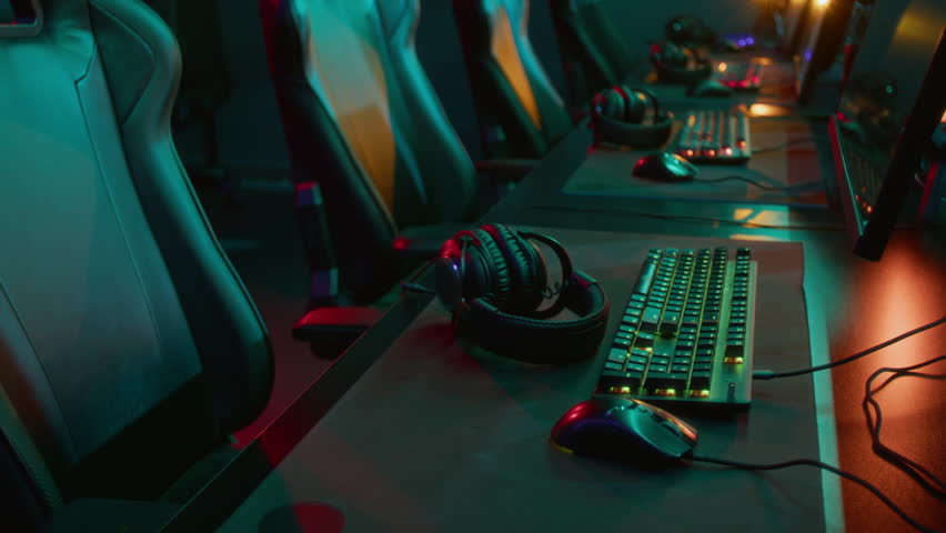 Medium no people shot of row of desks with computers, keyboards, mouses, headsets at online gaming cyberclub, in dim green and purple neon light before competition