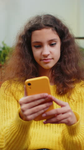Young teenager child girl texting share messages content on smartphone social media applications online watching relax movie. Female kid uses mobile phone at home in living room sits on sofa. Vertical