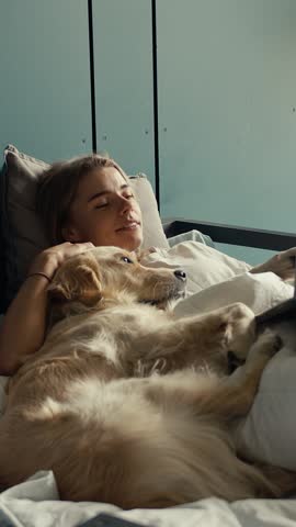 On a peaceful morning, a woman lounges in bed with her golden retriever, enjoying quality time together while watching entertaining content on her laptop