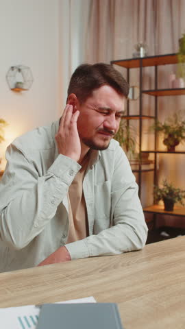 Sick young Caucasian man freelancer sitting at home office table feeling ear pain, unhappy irritated businessman or student. Furuncle, earache, shingles, eczema, ear disease, otitis concept. Vertical.