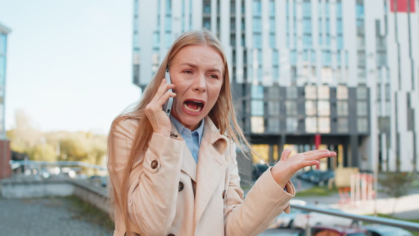 Angry mature businesswoman argues on smartphone while standing on downtown city street. Dissatisfied middle-aged woman girl in coat shouts into the mobile phone, expressing frustration and a problem.