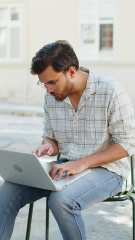 Handsome young man using laptop computer celebrating win good message news, lottery jackpot victory, giveaway online outdoors. Happy Indian guy tourist sitting in city cafe terrace. Town lifestyles.