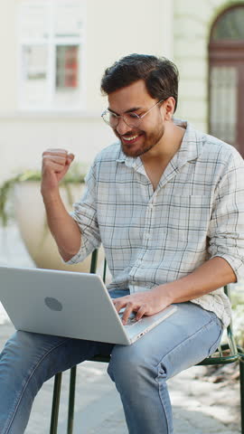 Handsome young man using laptop computer celebrating win good message news, lottery jackpot victory, giveaway online outdoors. Happy Indian guy tourist sitting in city cafe terrace. Town lifestyles.
