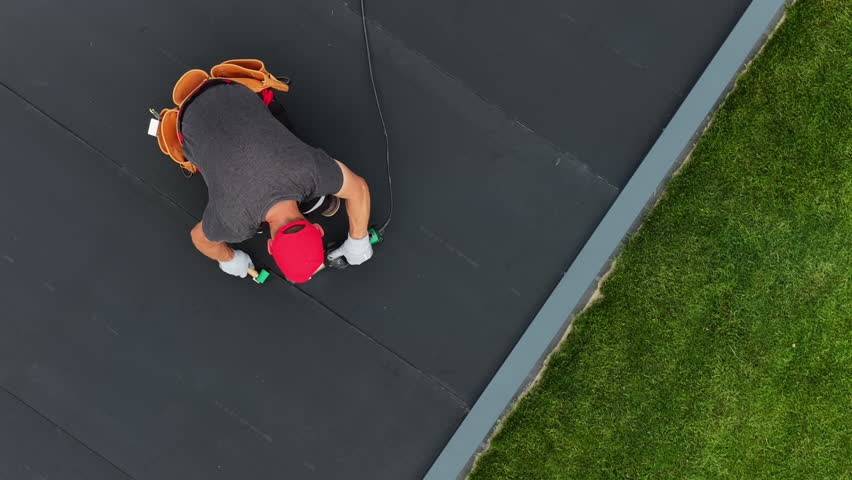 A worker repairs a EPDM roof using tools while on a ladder, with green grass visible below under clear skies. Safety equipment is clearly being used.