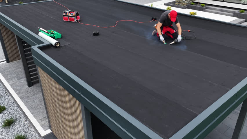 A worker carefully installs roofing material on a contemporary flat roof while using tools. The setting is bright with clean lines and modern architecture.