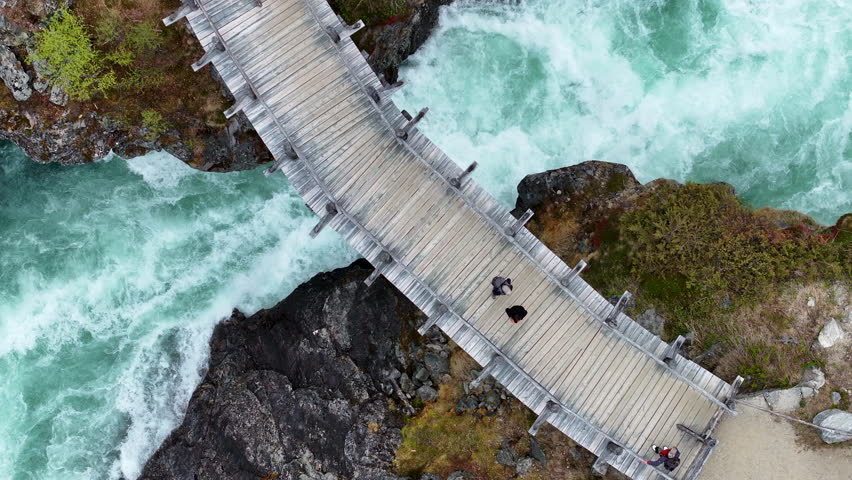 Explore a stunning wooden bridge spanning a vibrant river in Norway, surrounded by lush greenery and rocky terrain under bright sunlight.