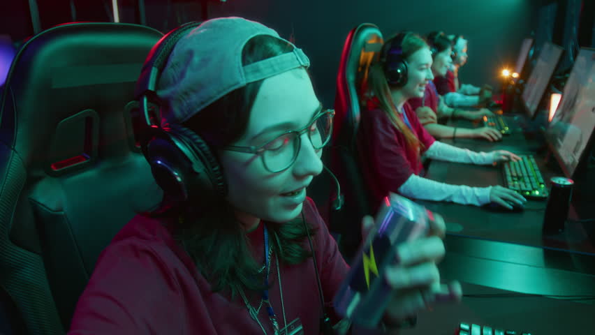 Medium UGC shot of joyous young Caucasian girl, wearing glasses and baseball cap, filming social media reel on smartphone with teammates, promoting energy drink, while playing video game tournament