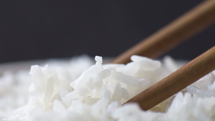 Wooden sticks taking pinch of hot fragrant rice. Macro shot. Bowl of freshly brewed Jasmine rice. Closeup view of long-grain variety of aromatic rice. Healthy eco food. Product of organic farming.