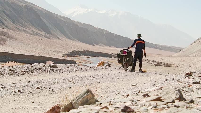 Static back view adventurous fit traveller cycle bicycle touring solo in remote high Pamir mountains early morning by river. Pamir highway Bartang valley