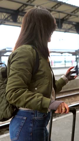 Female traveler pulling wheeled suitcase and wearing backpack, navigating train station while checking smartphone for travel details and destination information