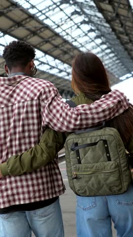 Multi ethnic couple walking arm in arm, sharing intimate moment inside modern train station, expressing joy and connection during travel journey