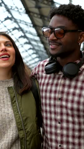 Multi ethnic couple walking side by side while chatting and smiling warmly inside bustling train station, sharing intimate moment during urban journey together