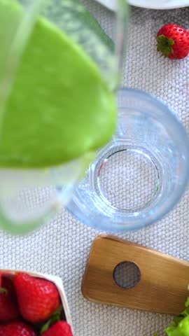 Vertical screen green smoothie pouring from blender into glass on table with fresh strawberries, avocado, and lettuce, promoting healthy eating and lifestyle