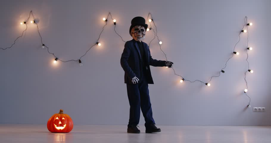 Young boy in scary mask and skeleton zombie costume having fun during Halloween holiday party. Cheerful kid raising up his legs and doing funny moves on garland lights background near pumpkin lantern.