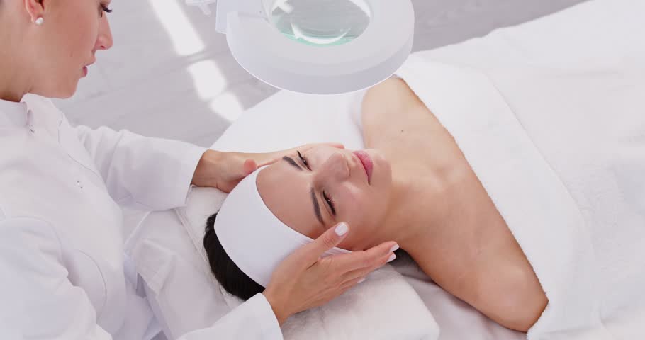 Professional cosmetologist is counseling her client on proper facial care techniques. In beauty salon woman lies on couch and cosmetologist stands next to her and talks about individual skin care tips