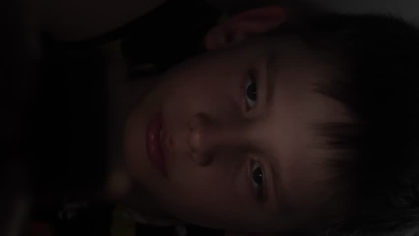 close up of a passionate Caucasian boy 11-12 years old using a gadget in the dark lying in bed, vertical video, light from the phone illuminates the child's face