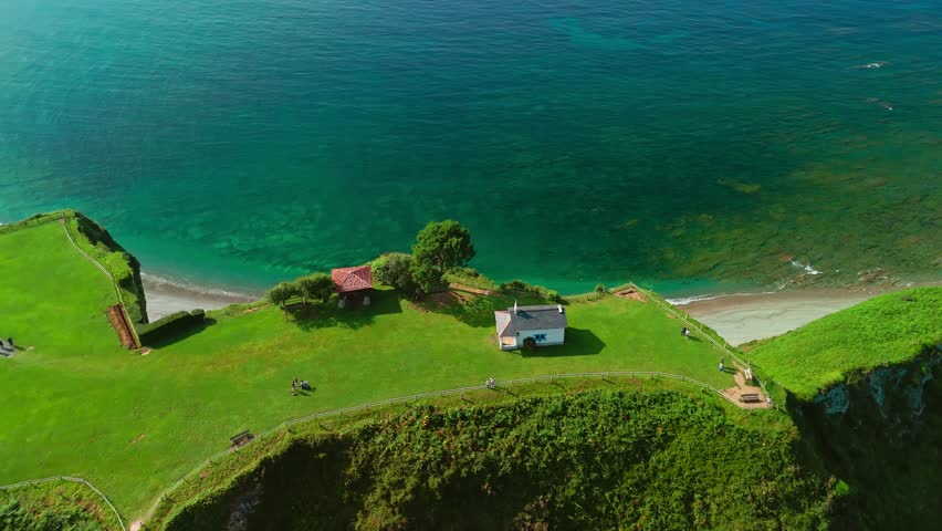 Aerial view of a small hermitage surrounded by stunning nature of Asturias in northern Spain. Ermita de la Regalina on the steep cliffs of the Cantabrian Sea coast. Beautiful Playa de Cadavedo beach