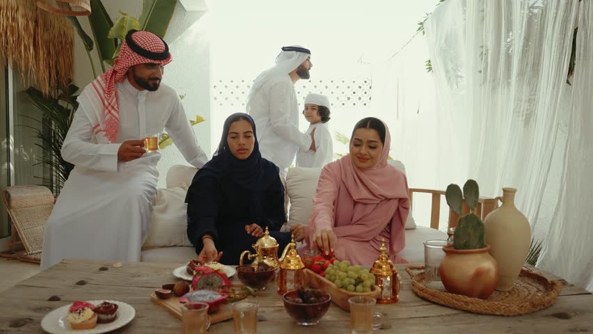 Emirati family wearing traditional clothing enjoying conversation and sharing diverse fruits and tea around wooden table in warm home interior