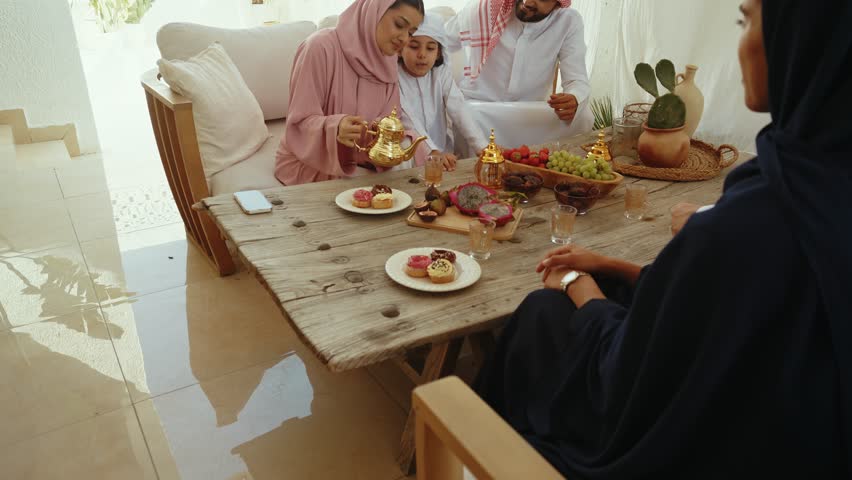 Emirati family wearing traditional clothing enjoying conversation and sharing diverse fruits and tea around wooden table in warm home interior