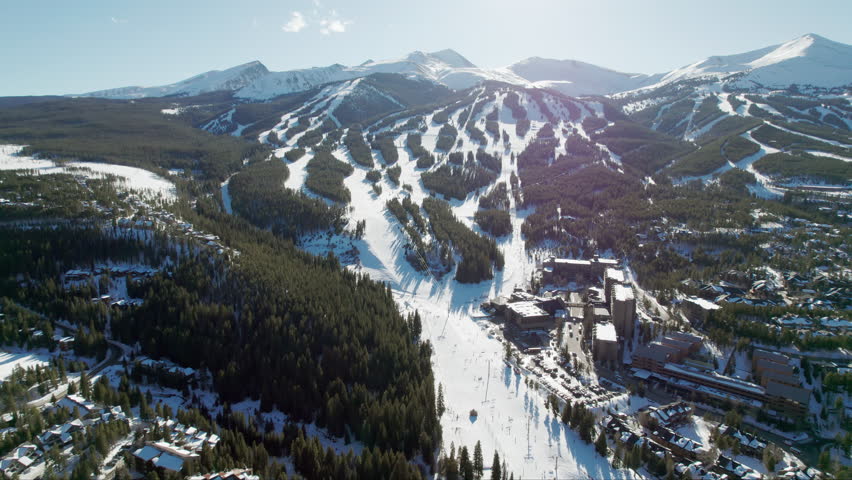 An aerial view approaching the popular Breckenridge ski resort in Colorado, with snow covered mountain peaks and high altitude trails full of winter skiers and snowboarders on a sunny winter day.