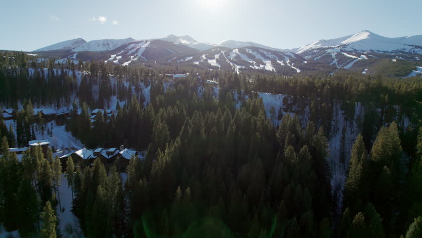 An epic aerial reveal of the popular Breckenridge ski resort in Colorado, with snow covered mountain peaks and high altitude trails full of winter skiers and snowboarders on a sunny winter day.