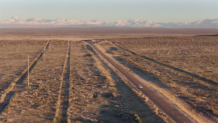 Route 40 cutting through Patagonian scrubland near Mendoza, Argentina, with vehicles heading south and the Andes rising in the distance under golden sunset light, drone panning, slow motion