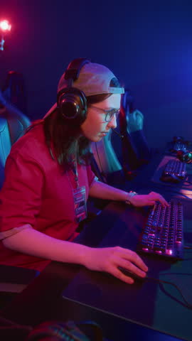 Vertical medium shot of geeky teenage Caucasian girl wearing glasses, baseball cap, headset sitting at computer in cyberclub, playing online video game, chewing gum, with focus and determination