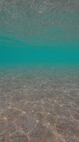 Vertical footage, Sun glare on the sea bottom covered with coarse sand in the evening light, Camera moving forwards approaching the shore over the seabed reflected in the surface of water