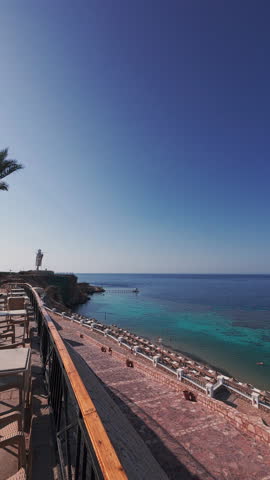 Sunny coastal resort by Red Sea. View from a stone terrace with sunbeds and railing overlooking calm turquoise waters and a coral reef. Bright sky and tranquil atmosphere. Vertical handheld footage