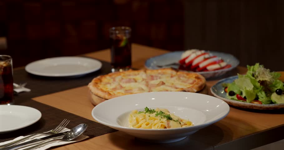Dinner setting restaurant table with pasta dish, pizza, salad, drinks. Waiter serving food, cozy ambiance, delicious meal, Italian dining experience.