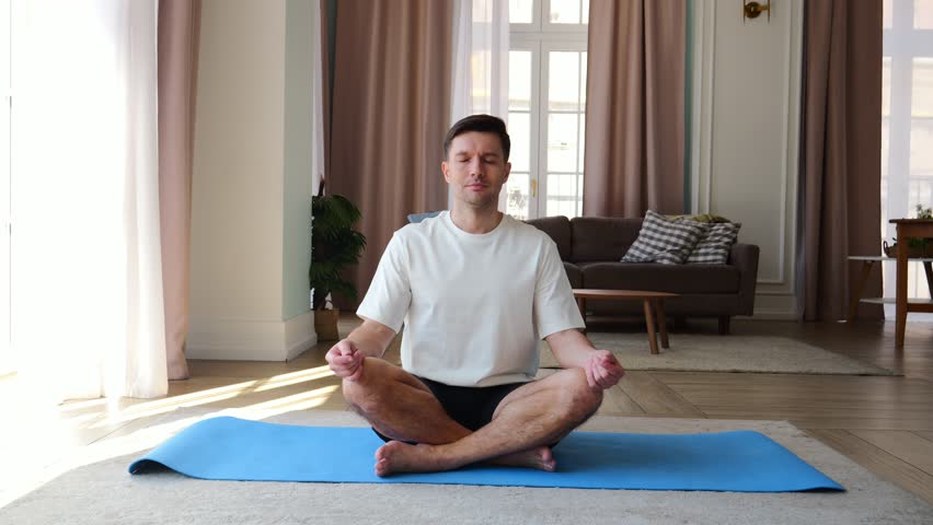 A man calmly meditates on a yoga mat, enjoying tranquility in a serene living space filled with soft sunlight.	
