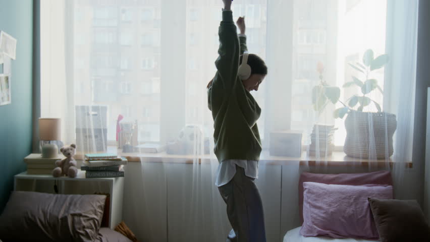 Wide angle shot of Caucasian teenage girl dancing with energy and enjoying music in dorm room while wearing big headphones backlit by window